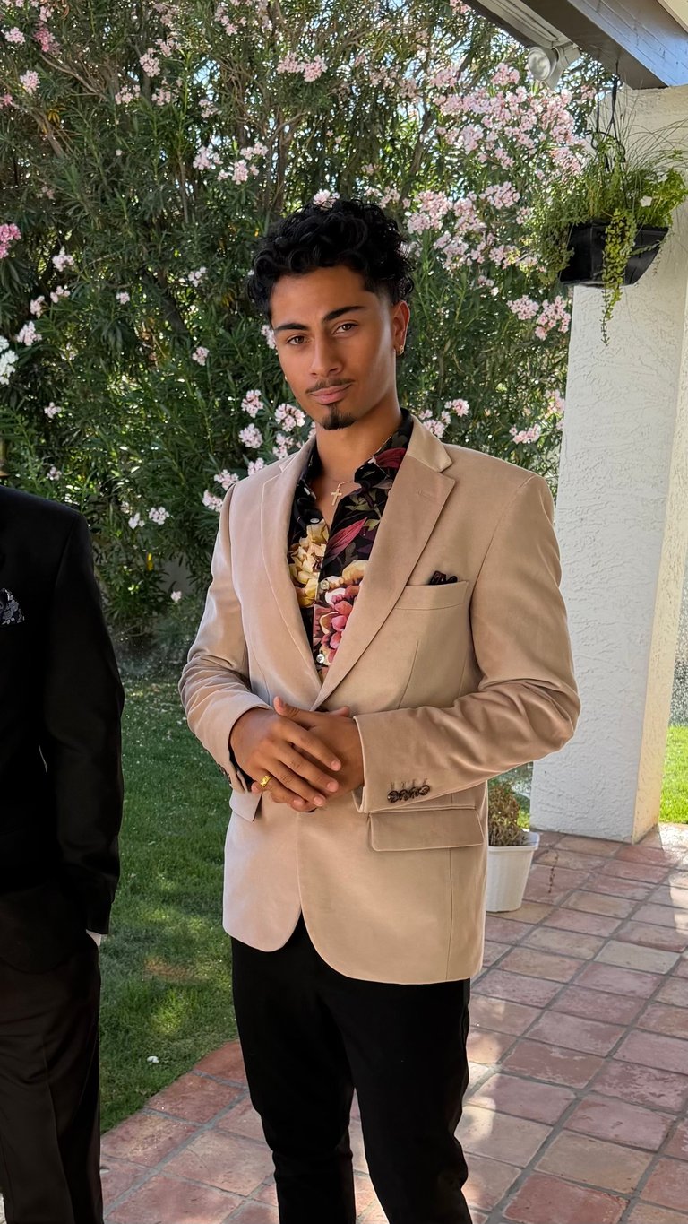 Man in beige blazer and black pants posing in front of lush green wall with pink flowers